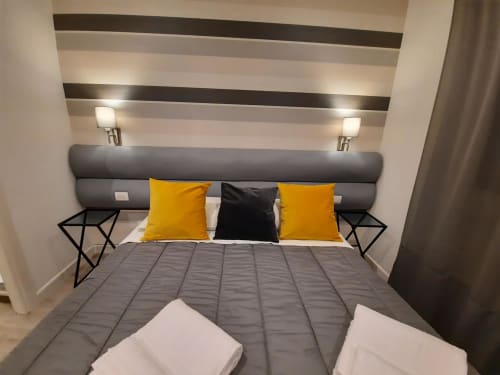 Camera moderna della Maritza Guest House Roma con letto grigio, cuscini gialli, testata rigata e illuminazione contemporanea
