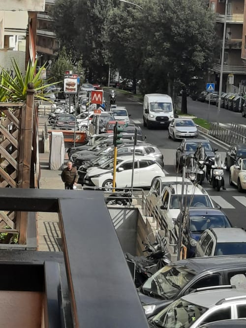 Vista della vivace strada romana dal balcone della Maritza Guest House con auto parcheggiate, scooter ed edifici urbani