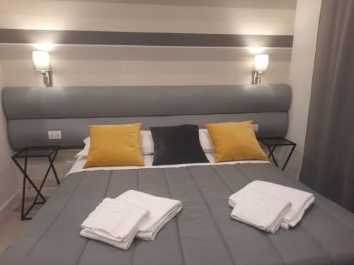 Camera moderna alla Maritza Guest House di Roma con letto imbottito grigio, cuscini gialli, asciugamani bianchi e illuminazione comodino
