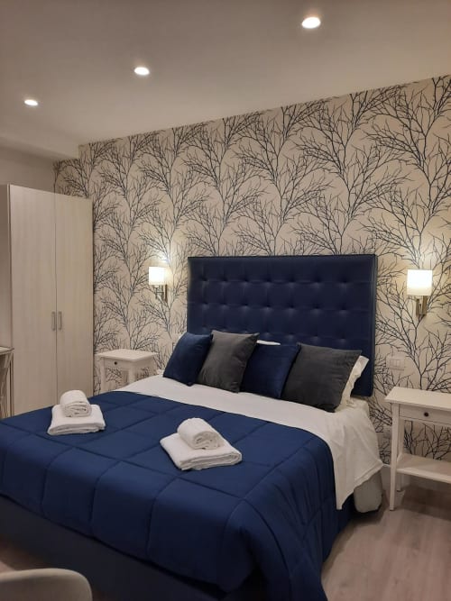 Camera moderna della Maritza Guest House Roma con letto imbottito blu scuro, carta da parati con motivi alberi e arredi contemporanei bianchi