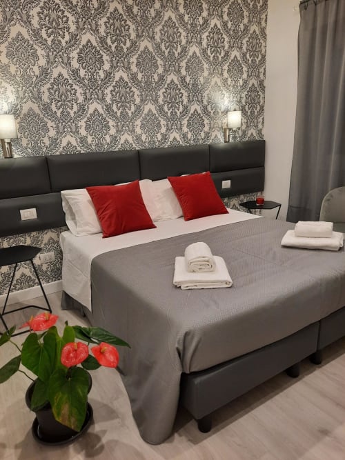Camera moderna della Maritza Guest House Roma con letto grigio, cuscini rossi, carta da parati damasco e fiori freschi