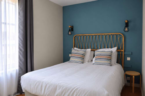 Chambre moderne à l'hôtel Au Bon Séjour avec mur d'accent bleu-vert, tête de lit en rotin, oreillers rayés et applique murale noire contemporaine