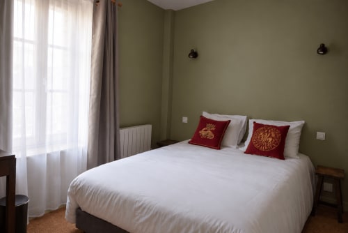 Chambre d'hôtel Au Bon Séjour à Sainte-Suzanne avec lit blanc, coussins rouges, murs olive et vue sur la ville