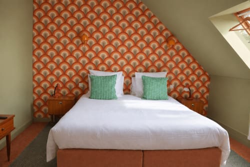 Chambre confortable à Au Bon Séjour avec papier peint orange à motifs, literie blanche, oreillers verts et tables de nuit en bois vintage