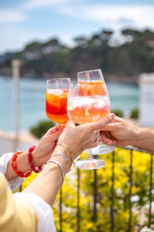 Dos personas brindando con cócteles Aperol Spritz frente a la costa de Llafranc en el Hotel La Muntanya