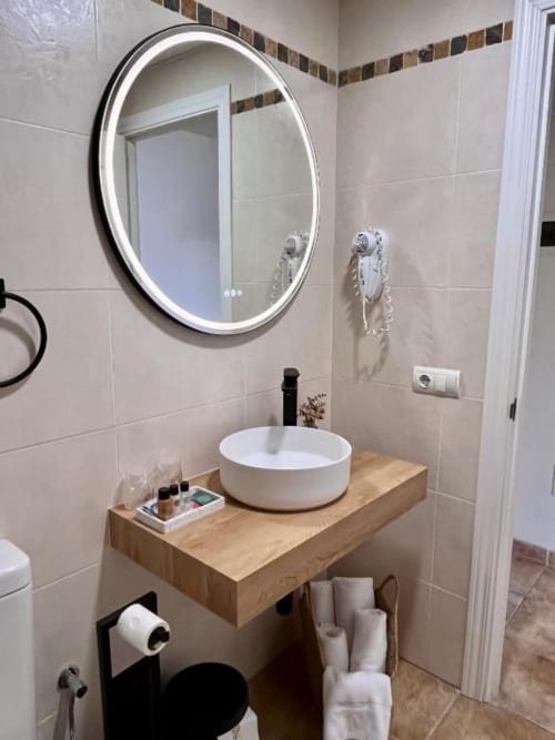 Baño moderno en la habitación Comfort del Hotel La Muntanya con espejo LED redondo, lavabo de cerámica blanca sobre tocador de madera, y