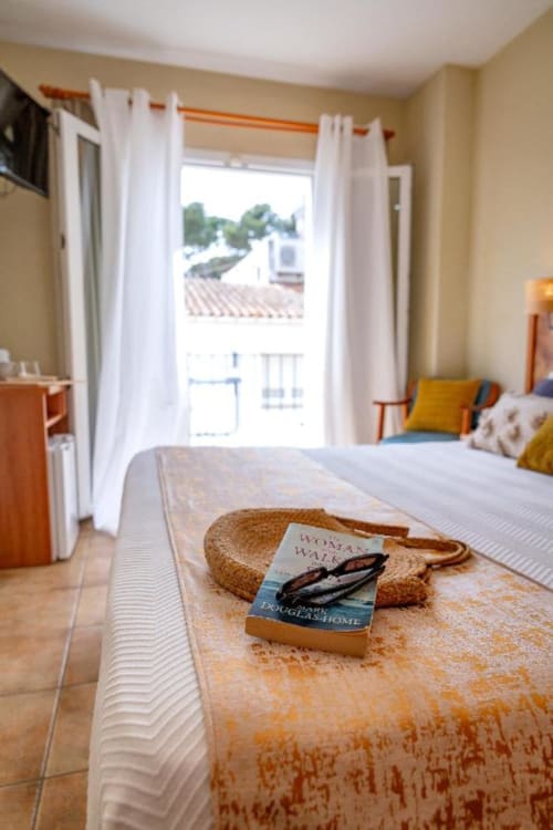Habitación luminosa en Hotel La Muntanya en Llafranc con libro sobre la cama y vistas desde el balcón a tejados y árboles