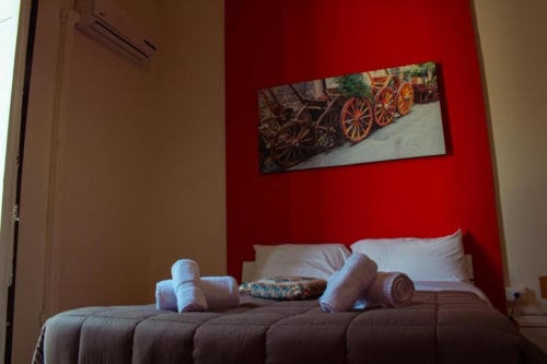 Luminosa camera tripla a Mizzica Rooms Palermo con parete accent rossa, letto confortevole, cuscini bianchi e decorativa ruota di carro