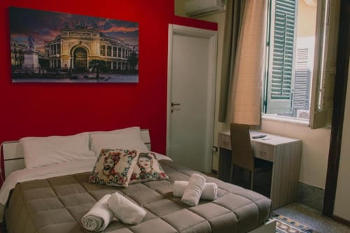 Camera da letto doppia luminosa a Mizzica Rooms a Palermo con pareti rosse, asciugamani arrotolati, cuscino colorato e poster teatrale