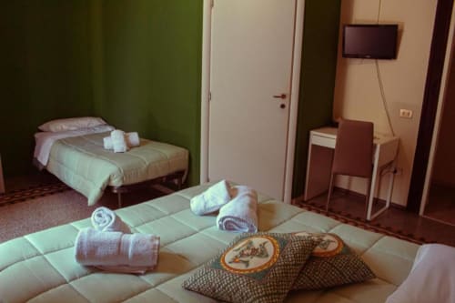 Camera tripla Mizzica Rooms Palermo: due letti, asciugamani arrotolati, cuscini decorativi, scrivania, TV, pareti verde salvia.