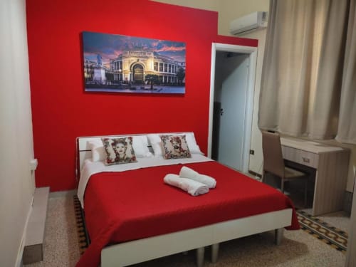 Camera da letto rosso brillante a Mizzica Rooms a Palermo con letto matrimoniale, cuscini bianchi, asciugamani arrotolati e skyline illuminato