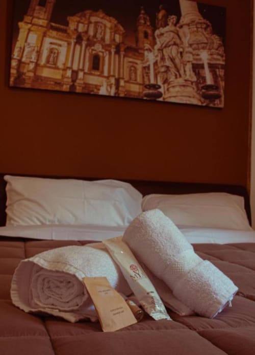 Asciugamani bianchi arrotolati con amenità omaggio sul letto di Mizzica Rooms a Palermo, con opere d'arte della città sulla parete