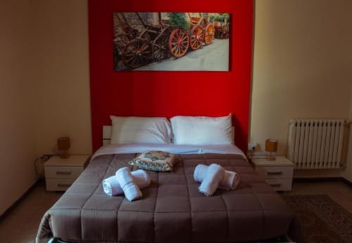 Camera tripla Mizzica Rooms Palermo: parete rossa, cuscini bianchi, asciugamani arrotolati e quadro carrello vintage