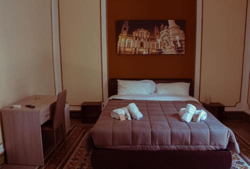 Camera quadrupla presso Mizzica Rooms a Palermo con biancheria da letto bordeaux, cuscini bianchi, asciugamani e opere d'arte dell'architettura siciliana sopra il