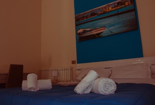 Asciugamani bianchi arrotolati sul letto della camera quadrupla con vista sulla città, con quadri raffiguranti barche e il waterfront di Palermo
