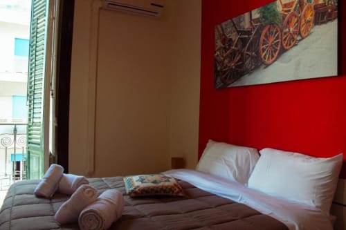 Luminosa camera tripla alle Mizzica Rooms di Palermo con parete accent rossa, biancheria bianca, accesso al balcone e vista sulla città