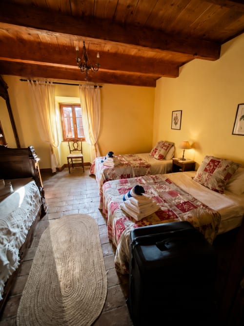 Rustic bedroom with exposed wooden beams, yellow walls, twin beds with red patterned quilts, and terracotta tile floor in a