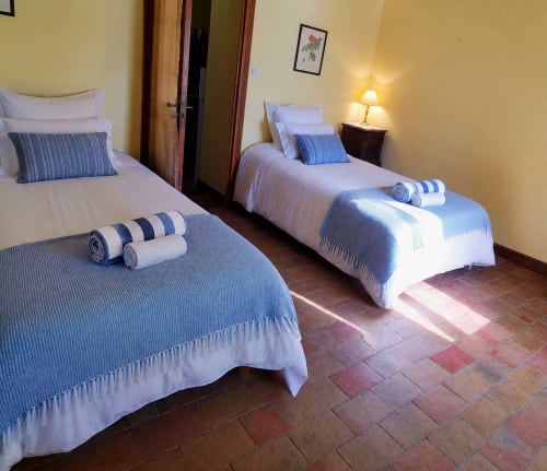 Twin beds with blue striped pillows in a bright cottage bedroom with terracotta floors and yellow walls at Mes Maisons in