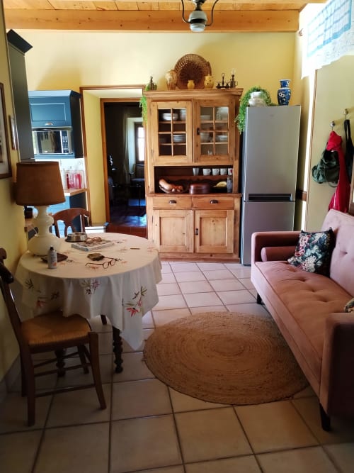 Cozy cottage kitchen-living area with yellow walls, wooden hutch, pink sofa, round jute rug, and dining table in French