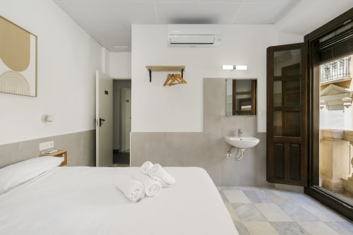 Moderna habitación del hotel Pensión Castil Granada con cama blanca, lavabo compartido y puertas de madera oscura hacia el balcón
