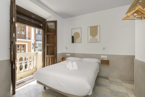 Habitación luminosa en Pensión Castil Granada con ropa de cama blanca, puerta de madera, vista de balcón a la ciudad y arte minimalista en las paredes