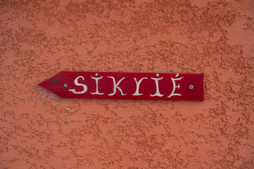 Panneau directionnel en bois rouge en forme de flèche affichant « SIKRIÉ » fixé sur un mur texturé de corail au gîte Le Nid De Bellemont, Trois-Rivières