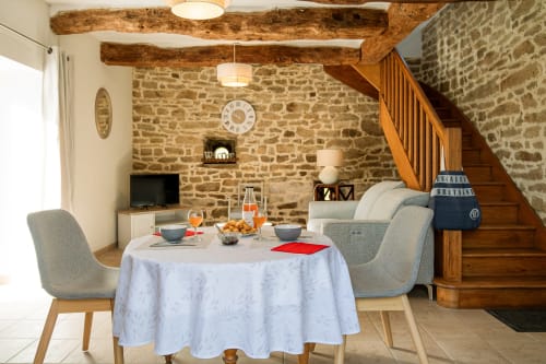 Salle à manger chaleureuse à la Ferme de Kervihern avec murs en pierre, poutres rustiques et table ronde dressée pour les hôtes.