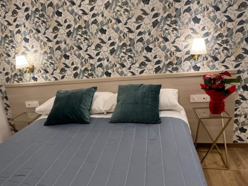 Camera da letto della Maritza Guest House a Roma con carta da parati decorata, coperta blu, cuscini verdi e fiori rossi sul comodino
