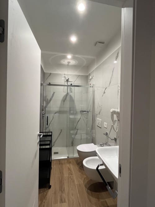 Bagno moderno nella Maritza Guest House Roma con doccia in vetro, sanitari bianchi e pavimento in legno