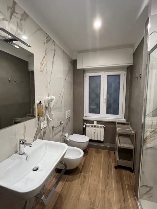 Bagno moderno della Maritza Guest House Roma con lavabo bianco, bidet, wc, piastrelle in marmo, pavimento in legno e finestra