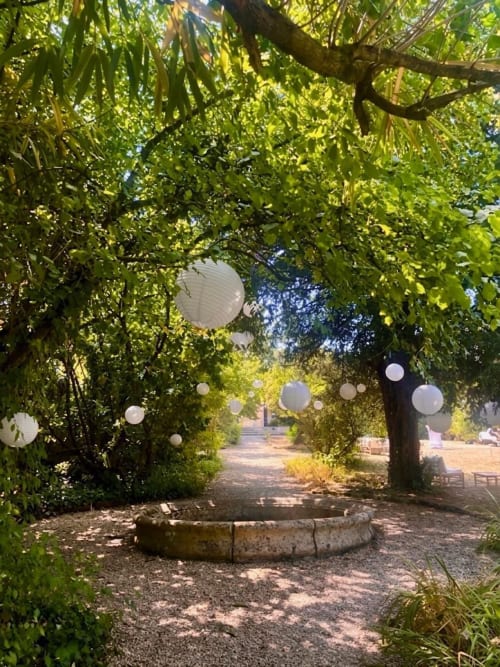 Allée enchantée du Domaine de Belisle avec lanternes suspendues, arbres luxuriants et coin de détente en pierre rustique