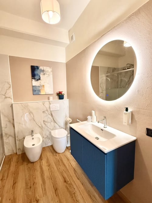 Bagno moderno dell'Hotel Il Mulino con specchio rotondo illuminato, vanità blu, dettagli in marmo, pavimento in legno e bianco