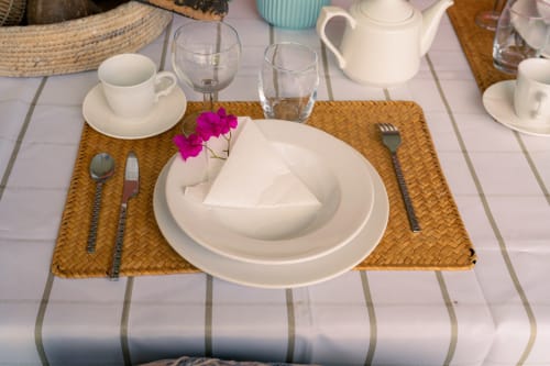 Mise de table élégante au Nid De Bellemont avec sets tressés, vaisselle blanche, fleurs roses et vue sur cottage bord de mer