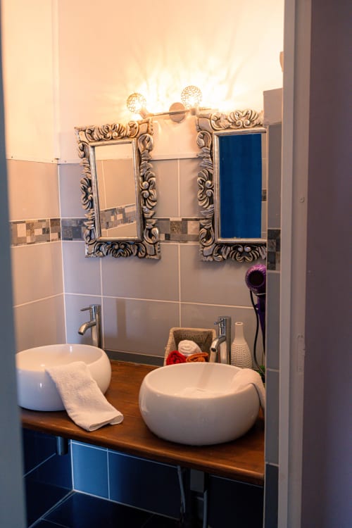 Salle de bains élégante avec deux vasques blanches, miroirs ornés et meubles bleus au gîte Le Nid De Bellemont