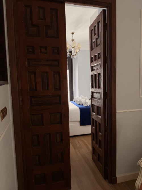 Puertas de madera oscura abriéndose al dormitorio de la Suite Giraldillo, Edificio San Esteban 16, Sevilla, con araña y toallas enrolladas
