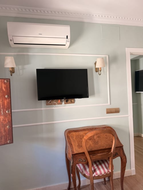 Habitación de hotel moderna en Sevilla con TV mural, zócalo blanco, silla antigua de madera y aire acondicionado split