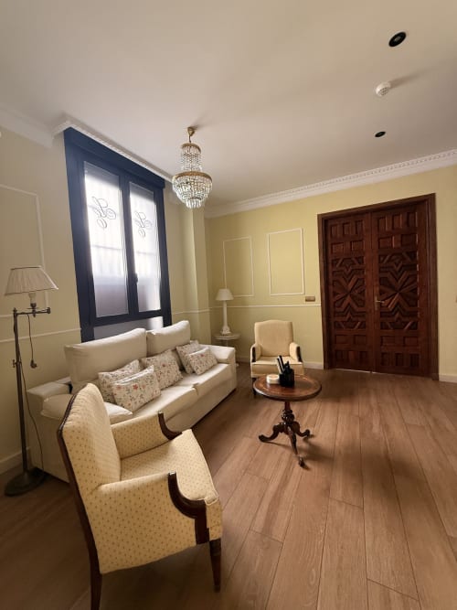 Elegante sala de estar de la suite en Edificio San Esteban 16 en Sevilla con sofá crema, puertas de madera, lámpara de araña y vistas a la ciudad.