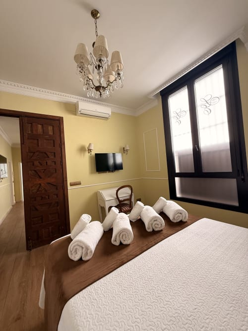 Habitación de suite del hotel Sevilla con toallas blancas enrolladas en la cama, paredes amarillas, puerta de madera oscura y ventanas con marco negro con vistas a la ciudad