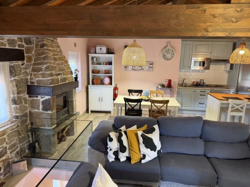 Acogedora sala de estar diáfana en casita rural asturiana con chimenea de piedra, sofá azul, mesa comedor y paredes rosa.