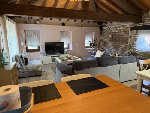 Salón moderno y comedor en propiedad rural asturiana con vigas de madera, chimenea, televisor y asientos cómodos