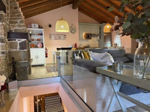 Sala de estar rústica moderna con chimenea de piedra, sofá gris, lámparas colgantes amarillas y cocina abierta en refugio rural asturiano