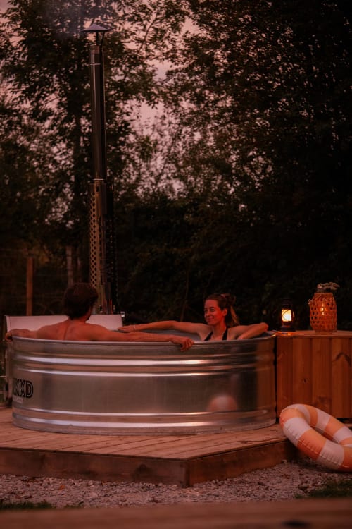 Deux hôtes se détendent dans un bain nordique à la chaumière Mesnil & Une Nuit, entourés d'arbres du jardin au crépuscule