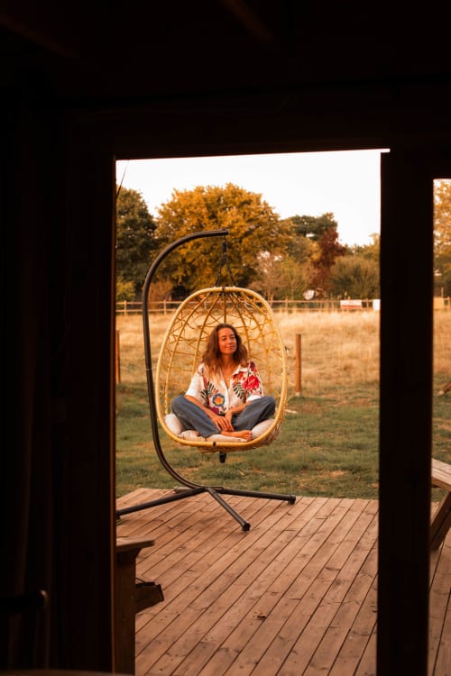 Femme se relaxant dans un fauteuil œuf suspendu sur la terrasse en bois surplombant le jardin du gîte Mesnil & Une Nuit, France