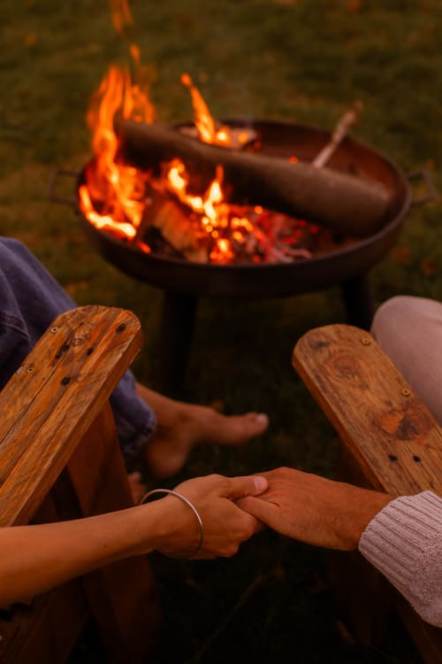 Couple se tenant la main près d'un foyer au gîte Mesnil & Une Nuit, avec des chaises Adirondack et les flammes qui dansent