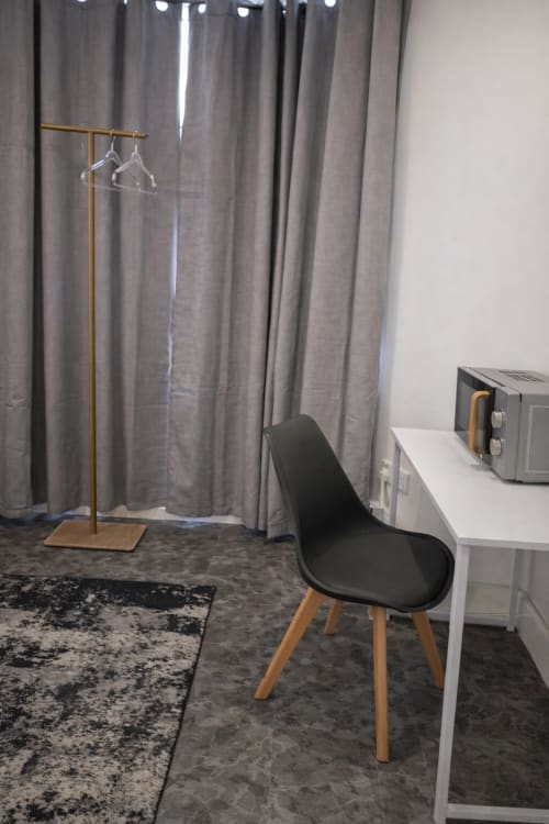 Modern hotel room at La Rose Serenity Residence in London with black chair, white desk, clothing rack, and grey curtains