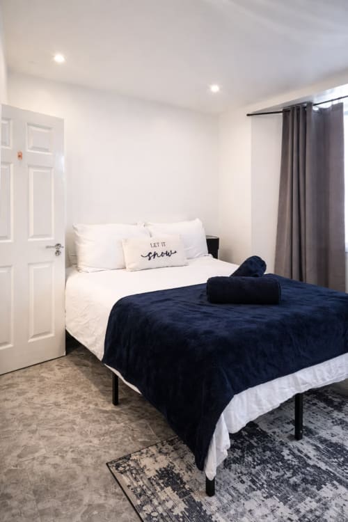 Modern bedroom at La Rose Serenity Residence in London with queen bed, navy blanket, white pillows, and grey curtains