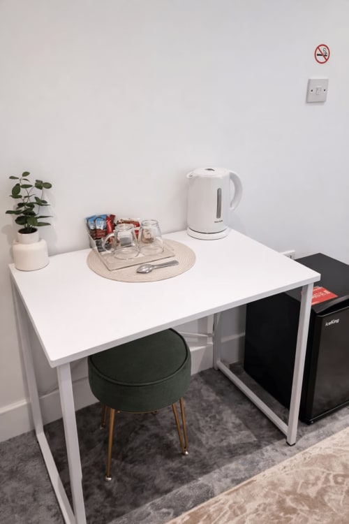 Compact hotel desk in London room with kettle, glasses, plant, and green stool
