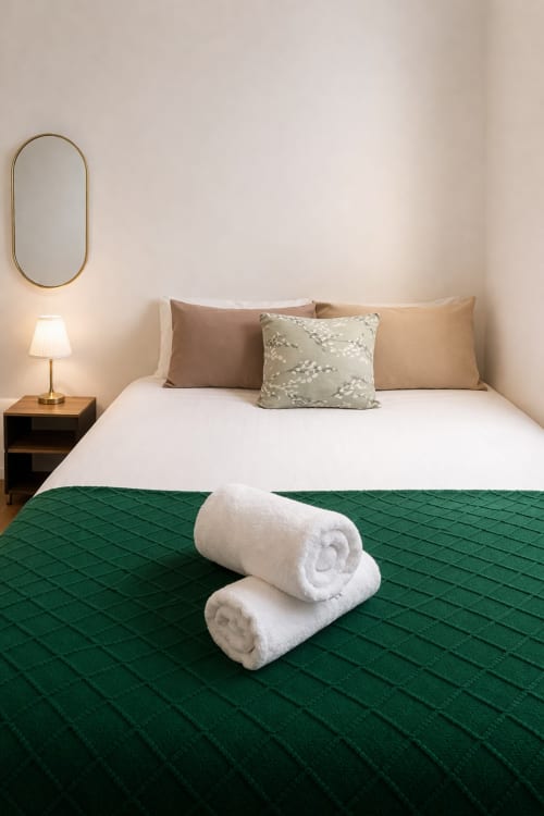 Elegant London B&B bedroom with white towels on green quilted throw, beige pillows, brass mirror, and bedside lamp