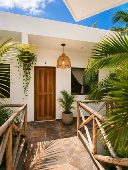 Entrada de la Suite 14 en Hotel Holbox Centro con puerta de madera, lámpara colgante tejida y plantas tropicales bajo cielo azul