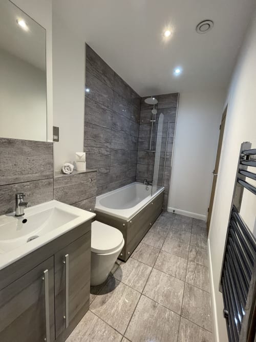 Modern bathroom in Empire Serviced Apartments Leicester, featuring gray tiled walls, white sink, toilet, bathtub with shower